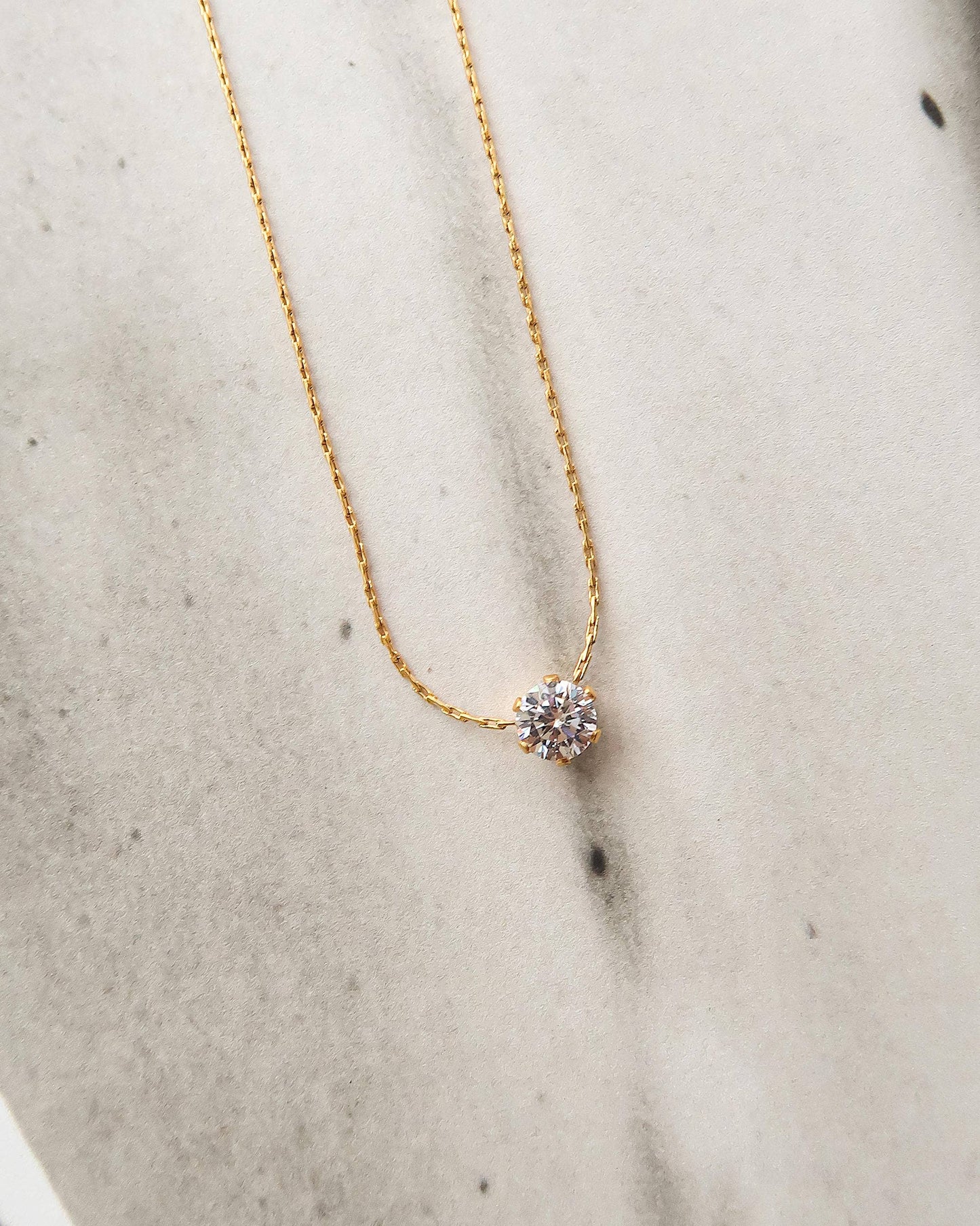 Close-up of the Serein Necklace, a dainty 18K gold-plated chain featuring a small round cubic zirconia pendant, photographed on a soft neutral display.