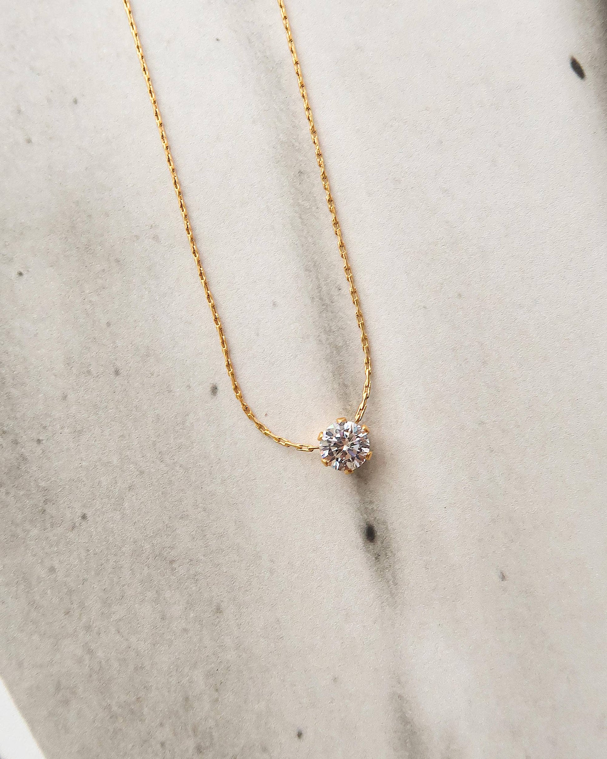 Close-up of the Serein Necklace, a dainty 18K gold-plated chain featuring a small round cubic zirconia pendant, photographed on a soft neutral display.
